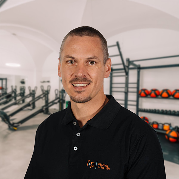 Ein lächelnder Mitarbeiter von f+p Gesund Bewegen steht im Trainingsraum vor Fitnessgeräten und trägt ein schwarzes Poloshirt.