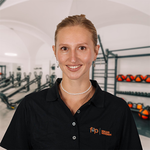 Melanie von f+p Gesund Bewegen lächelt in einem schwarzen Poloshirt mit Firmenlogo vor Trainingsgeräten und Medizinbällen.