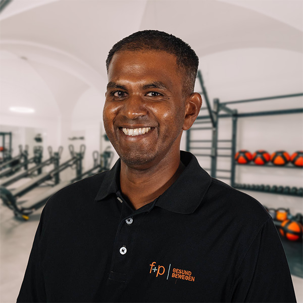 Der lächelnde Mitarbeiter Michael von f+p Gesund Bewegen steht im Trainingsraum zwischen Fitnessgeräten und Medizinbällen.