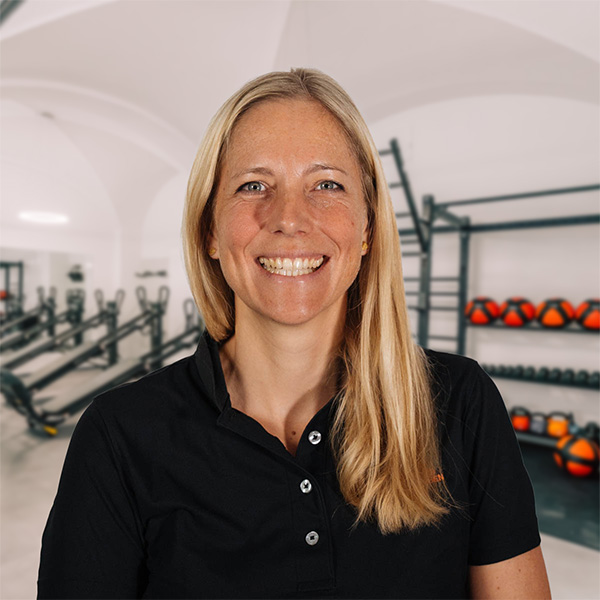 MoHa Die lächelnde Monika trägt ein schwarzes Polohemd und steht vor Trainingsgeräten und Medizinbällen im Therapie- und Fitnessbereich.