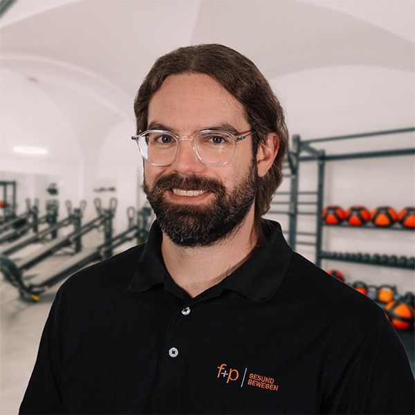 Ein lächelnder Physiotherapeut mit Brille und dunklem Bart steht im Trainingsbereich von f+p Gesund Bewegen in Kempten.